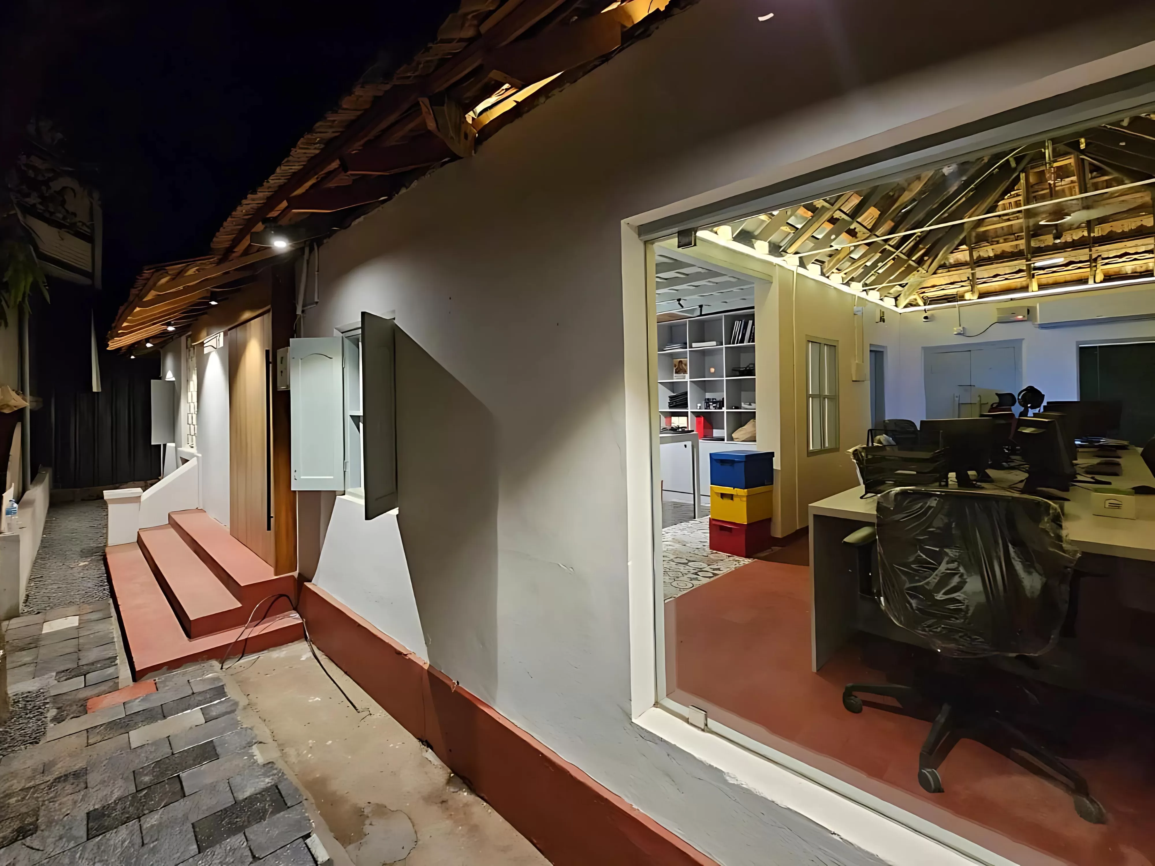 Night view of Living Impressions' office, featuring a blend of traditional and modern architectural design in Kochi. The office showcases an exposed wooden beam roof, large glass windows, and a well-lit minimalist entrance with sleek steps, highlighting traditional and modern architectural design in Kochi.